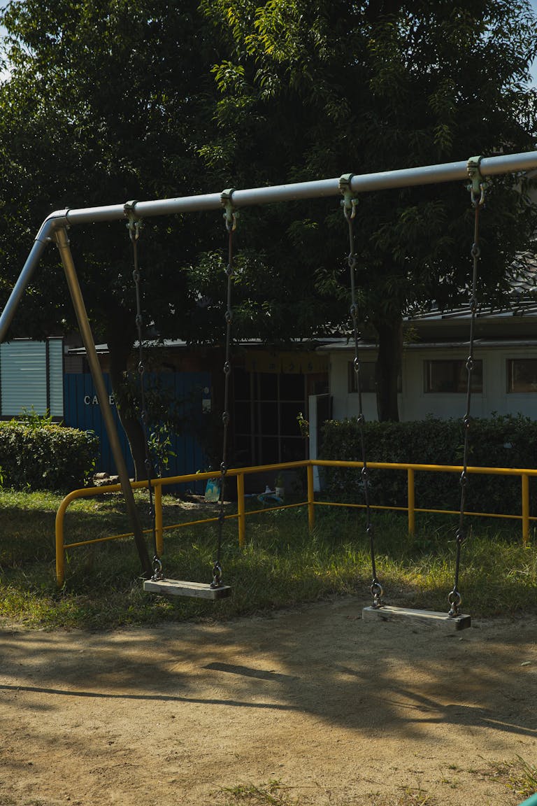 Metal empty swings on playground in yard among green trees on sunny summer day