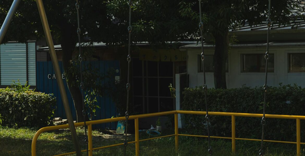 Metal empty swings on playground in yard among green trees on sunny summer day