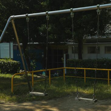 Metal empty swings on playground in yard among green trees on sunny summer day